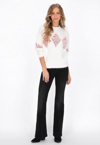 Weißer Strickpullover mit rotem Rautenmuster, langen Ärmeln und gerippten Bündchen, kombiniert mit schwarzen Schlagjeans. Einfarbige schwarze Knöchelstiefel.