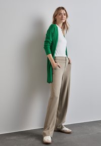 Un long cardigan vert sur un t-shirt blanc, un pantalon beige à jambes larges et des baskets claires. Textures lisses, coupe décontractée et poches pratiques.