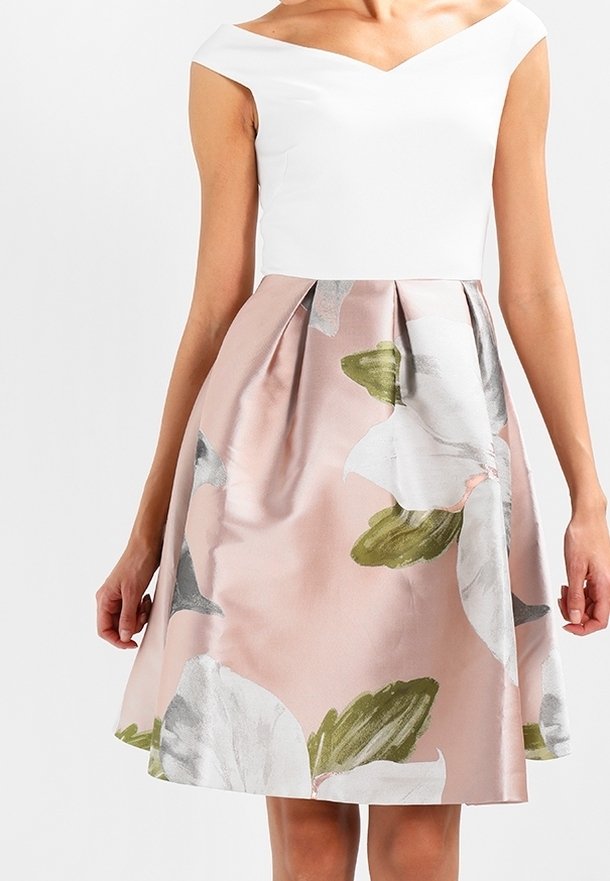 Femme portant une robe blanche épaules dénudées avec une jupe rose ornée de grandes fleurs blanches et de feuilles vertes.