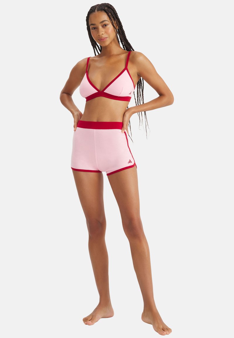 Femme aux cheveux tressés, debout pieds nus, portant un brassière de sport rose et un short avec bordure rouge, posant devant un fond uni.