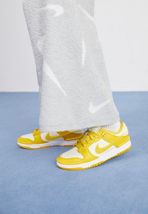 Persona con calcetines blancos y zapatillas Nike amarillas y blancas, de pie sobre un suelo azul con una falda gris texturizada que cubre parte de las piernas.