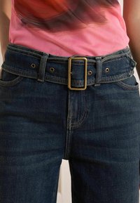 Tumed denim teksad keskmise tõusuga, vaskse vöölukuga, viie taskuga ja peente õmblusdetailidega. Roosa topp on nähtav vöö kohal.