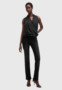 Svart ermeløs bluse med blondedetaljer, drapert design, kombinert med tettsittende svarte jeans og åpne svarte hæler, står foran en ensfarget bakgrunn.