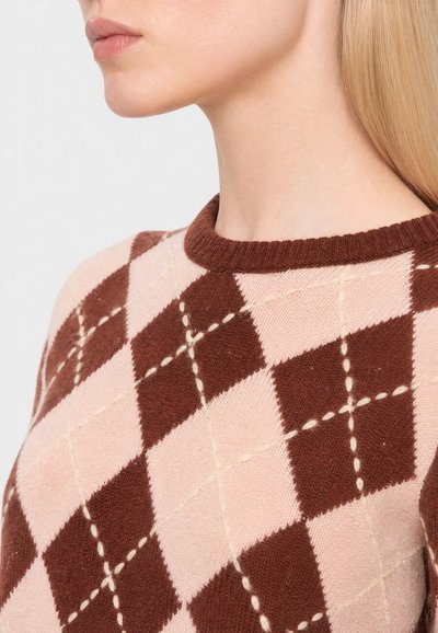 Femme portant un pull à motif losanges marron et rose, recadré pour montrer le cou, le menton et une partie des cheveux blonds mi-longs.