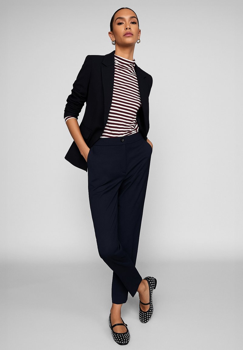 Pantaloni sartoriali blu navy, blazer nero, top a strisce bordeaux e bianche, e ballerine nere a pois. Tessuto liscio, design aderente e linee pulite.