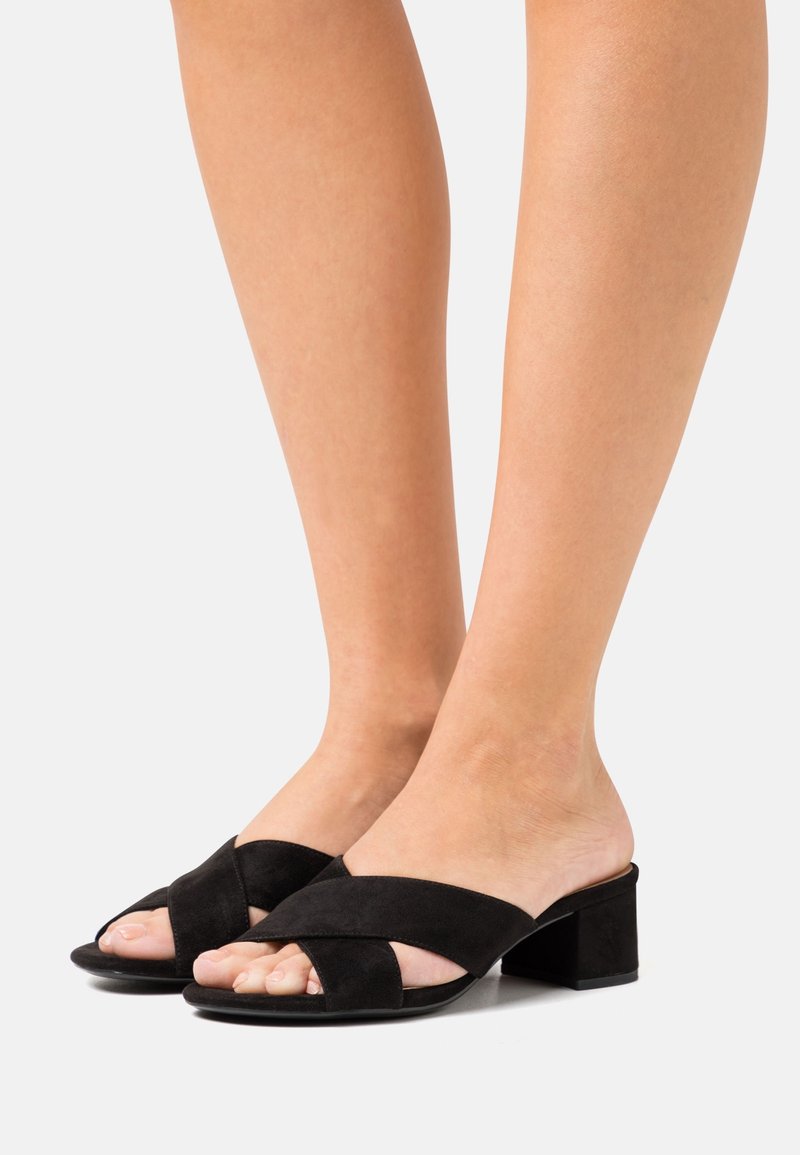 Pieds portant des sandales à talons blocs ouvertes au bout en suède noir avec de larges brides croisées, présentés sur un fond clair uni.
