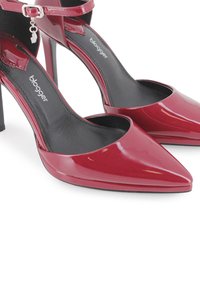 Zapatos de tacón alto de charol rojo con punta afilada, correa en el tobillo y un forro interior negro, que cuentan con un pequeño encanto decorativo en la correa.