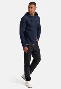 Marineblauer Hoodie mit geripptem Textil und Kordelzug, kombiniert mit schwarzen Jeans und weißen Turnschuhen. Einfaches, lässiges Design.