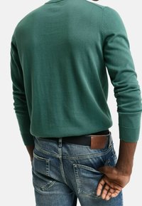 Grüner Pullover mit gerippten Bündchen und Kragen, kombiniert mit blauen Jeans, die mehrere Taschen und einen braunen Ledergürtel haben.