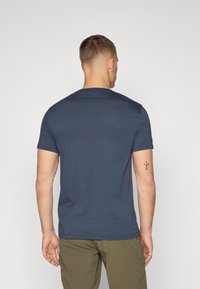 Námornícky modré tričko s krátkym rukávom a okrúhlym výstrihom, vyrobené z mäkkej tkaniny. Viditeľné švy na leme a rukávoch, nosené s olivovozelenými nohavicami.