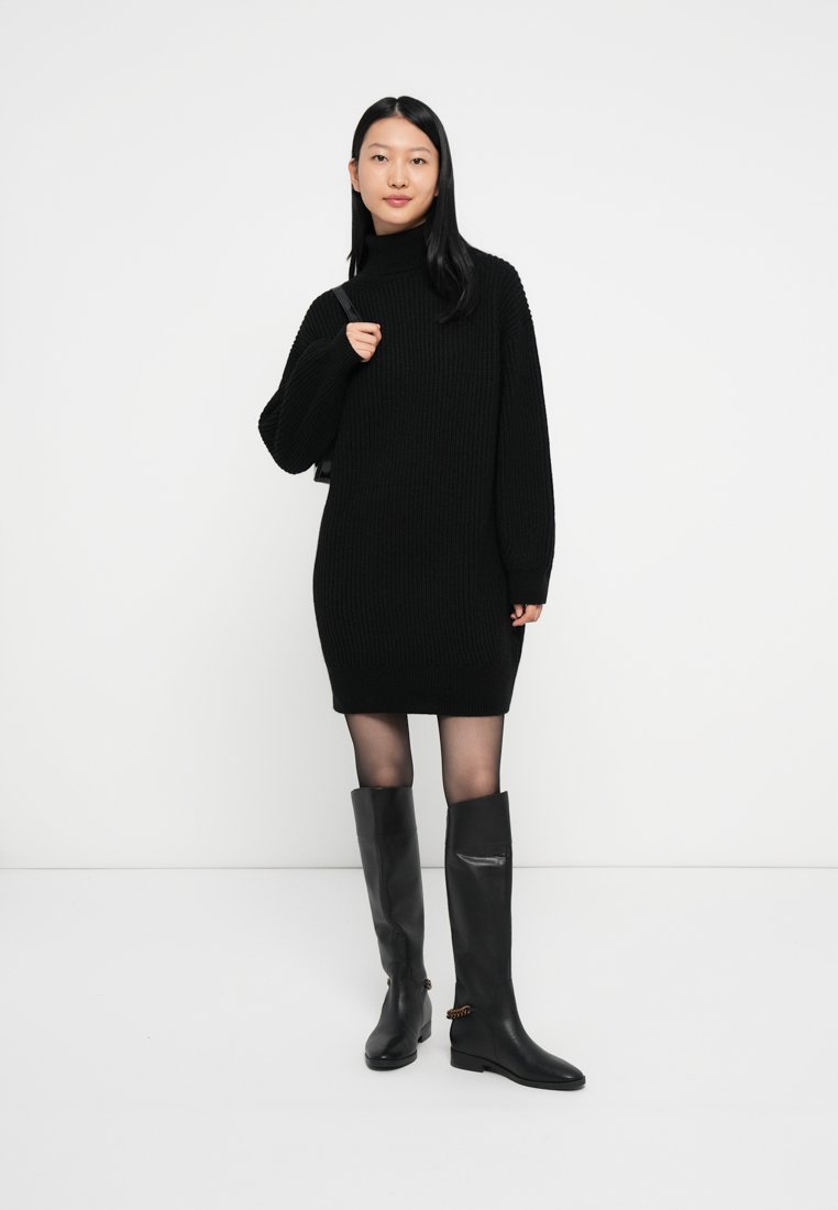 Robe pull côtelée noire avec col montant, associée à des bottes noires moulantes jusqu'aux genoux avec un accent décoratif. Texture lisse et coupe ajustée.
