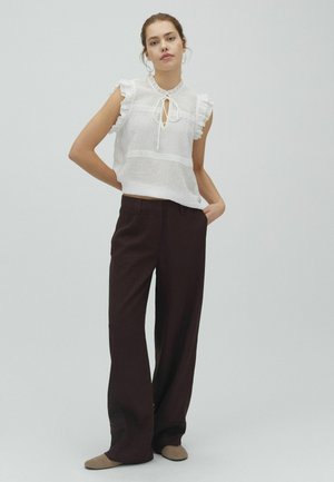 Kobieta z włosami związanymi do tyłu, nosząca duże kolczyki koła, białą, bezrękawną bluzkę z falbankami, ciemne szerokie spodnie i beżowe buty, stojąca z rękami w kieszeniach.