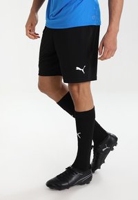 Schwarze Sportshorts mit weißem Puma-Logo, kombiniert mit schwarzen, knielangen Socken mit weißem Logo. Schwarze Fußballschuhe mit strukturiertem Design.