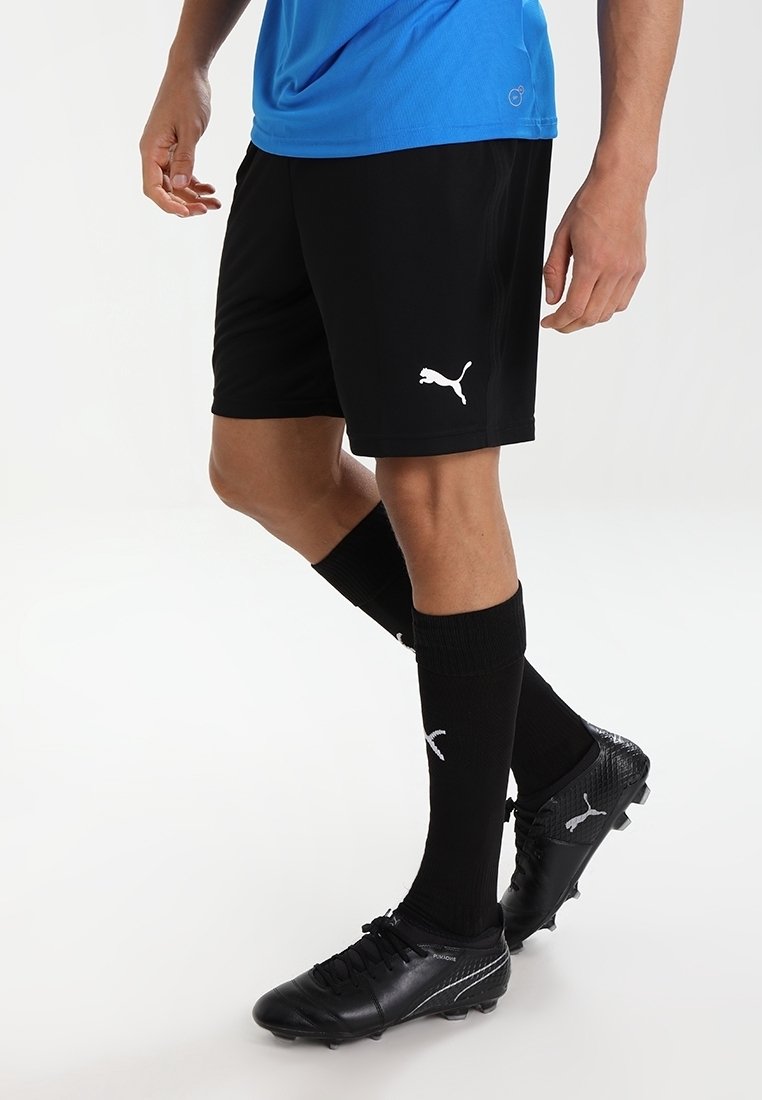 Schwarze Sportshorts mit weißem Puma-Logo, kombiniert mit schwarzen, knielangen Socken mit weißem Logo. Schwarze Fußballschuhe mit strukturiertem Design.