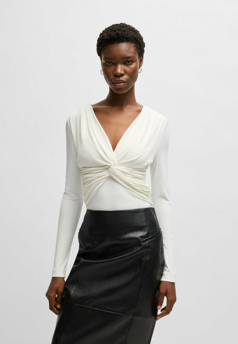 Top bianco a maniche lunghe con un design frontale annodato abbinato a una gonna in pelle nera a vita alta caratterizzata da una texture liscia e tasche sottili.