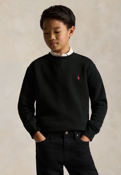 Junge trägt schwarzen Sweatshirt mit rotem Logo und schwarze Hose, steht mit Händen in den Taschen vor einfarbigem Hintergrund.