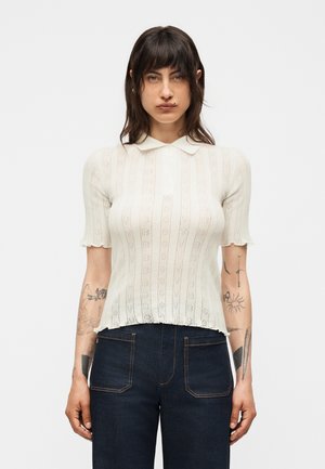 Kvinde med skulderlangt mørkt hår bærer en hvid striktop med korte ærmer og krave samt mørkeblå højtaljede jeans, der viser armtatoveringer.