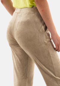 Pantalones de ante beige con una textura suave. Cuentan con bolsillos laterales y una silueta ajustada, mostrados desde atrás con una blusa verde vibrante.