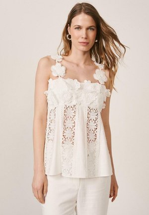 Mujer con camiseta blanca sin mangas con patrones de encaje floral y tirantes en forma de flores, combinada con pantalones blancos sobre fondo liso.