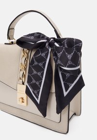 Beige handväska med en texturerad yta, prydd med ett guld kedjehandtag och en svart mönstrad scarf knuten ovanpå, accentuerad med vita detaljer.