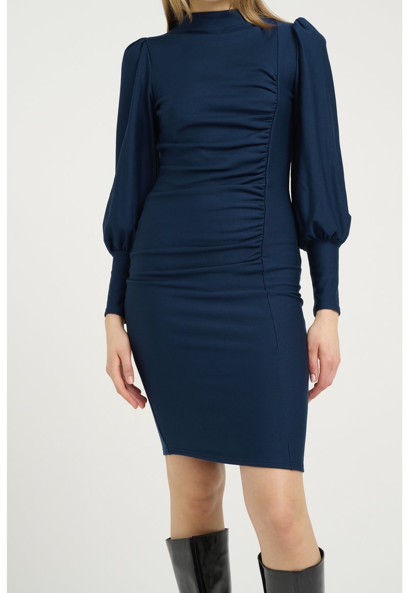 Robe ajustée bleu marine avec détail froncé, manches longues bouffantes, col montant, portée avec des bottes noires montantes au genou.