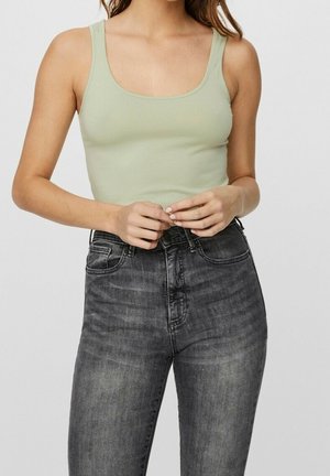 Vrouw die een lichtgroen mouwloos topje en hooggetailleerde vervaagde zwarte spijkerbroek draagt, handen samengevouwen ter hoogte van de taille, effen lichte achtergrond.