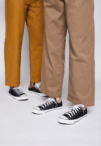Deux personnes portant des baskets noires et blanches avec des lacets blancs, l'une en pantalon jaune moutarde et l'autre en pantalon large marron clair.