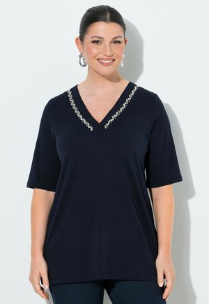 Uśmiechnięta kobieta o ciemnych włosach związanych w niski kok, nosząca srebrne kolczyki koła i granatowy top z dekoltem w serek zdobiony kryształkami.