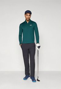 Camisola polo verde de mangas compridas com um logótipo, combinada com calças escuras. Usada com boné azul-marinho e luva branca, segurando um taco de golfe. Ténis atléticos azuis.