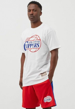 Uomo che indossa una maglietta bianca dei Los Angeles Clippers e pantaloncini da basket rossi in piedi con le mani in tasca.