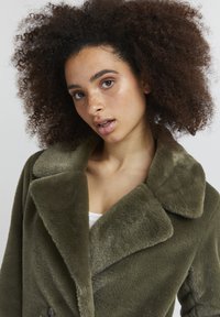 Faux fur olivengrøn frakke med bred krave, dobbeltradet knaplukning og en blød, struktureret overflade. En subtil glans forbedrer det samlede look.