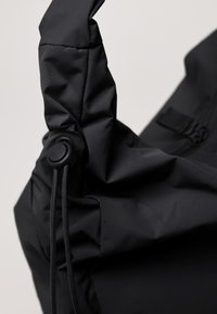 Schwarze Stofftasche mit strukturierter Oberfläche, ausgestattet mit einem genähten Griff und einer Zugkordelverstellung. Auffälliges Knopfdetail zum Verschließen.