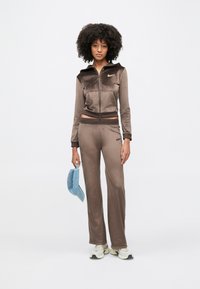 Veste zippée en velours marron avec capuche, assortie à un pantalon assorti. Les deux portent un logo. Le mannequin tient une casquette bleu clair.