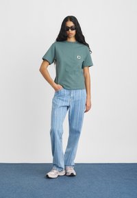 Roheline lühikeste varrukatega T-särk rinnataskuga, millele on sobitatud helesinised triibulised teksad. Modell kannab päikeseprille ja heledaid halli värvi tossusid.