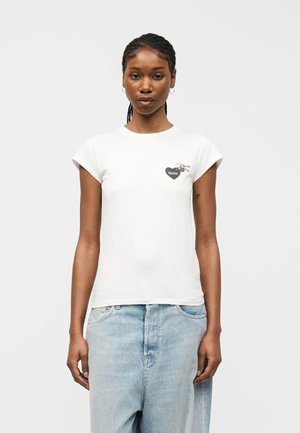 Młoda kobieta ubrana w białą, dopasowaną koszulkę z krótkim rękawem i małym czarnym logo w kształcie serca oraz w jasnoniebieskie, luźne dżinsy, na tle jednolitego koloru.