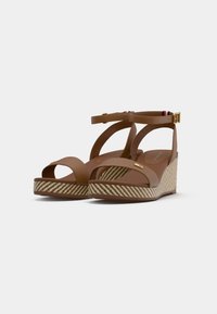 Brown wedge sandals with crisscross straps, a textured striped platform, and gold hardware accents. Adjustable ankle strap for fit.