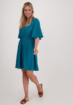Teal, mouwloze jurk met een V-hals en een tunneltaille, voorzien van een uitlopende rok en een gestructureerde stof. Bruine sandalen maken de look compleet.