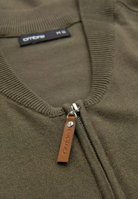 Pull zippé vert olive avec un col côtelé, doté d'une fermeture éclair en métal et d'une étiquette en cuir marron embossée avec le nom de la marque.