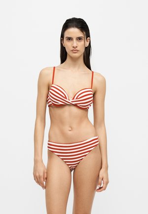 Femme aux longs cheveux foncés portant un bikini rayé rouge et blanc avec des fines bretelles sur un fond blanc uni.