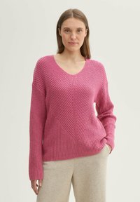 Maglione rosa a maglia con scollo a V, motivo a treccia e consistenza a costine; presenta maniche larghe e una vestibilità rilassata. Abbinato a pantaloni grigi chiari.