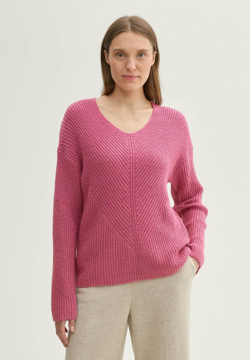 Maglione rosa a maglia con scollo a V, motivo a treccia e consistenza a costine; presenta maniche larghe e una vestibilità rilassata. Abbinato a pantaloni grigi chiari.