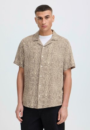 Jeune homme aux cheveux courts portant une chemise à manches courtes beige à motifs floraux par-dessus un T-shirt blanc et un pantalon noir, fond neutre.