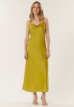 Jeune femme aux cheveux lâchés portant une robe nuisette en satin jaune moutarde à fines bretelles et des sandales à talons hauts dorées, fond neutre.