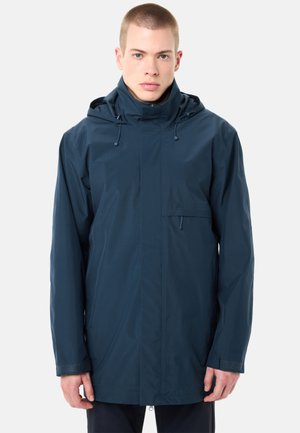 Junger Mann trägt marineblaue, kapuzenartige Regenjacke mit Reißverschluss und Kordelzug, steht vor einfachem, hellem Hintergrund.