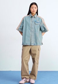 Mladá žena stojí proti světle modré pozadí, oblečená v džínové oversized košili, béžových cargo kalhotách a růžových teniskách.
