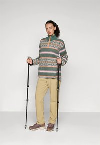 Vihreän, vaaleanpunaisen ja oranssin geometrisin kuvioin varustettu fleecetakki. Yhdistetty beigeihin housuihin ja ruskeisiin vaelluskenkiin, kädessä vaellussauvat.