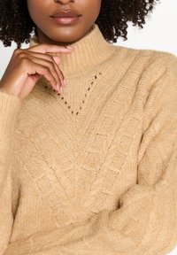 Pull beige en maille avec un col haut, présentant un motif chevron texturé et des poignets côtelés. Tissu en maille épaisse et douce.