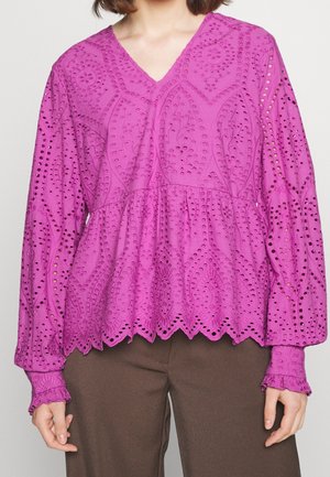 Femme portant un chemisier violet à manches longues avec une broderie ajourée complexe et un ourlet festonné, associé à un pantalon marron.