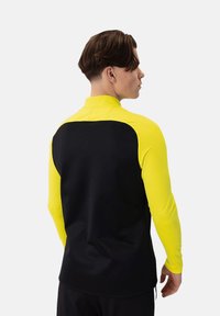 Maglietta sportiva a maniche lunghe con un design aderente, schiena nera e maniche gialle brillanti. Realizzata in tessuto elasticizzato con colletto alto.