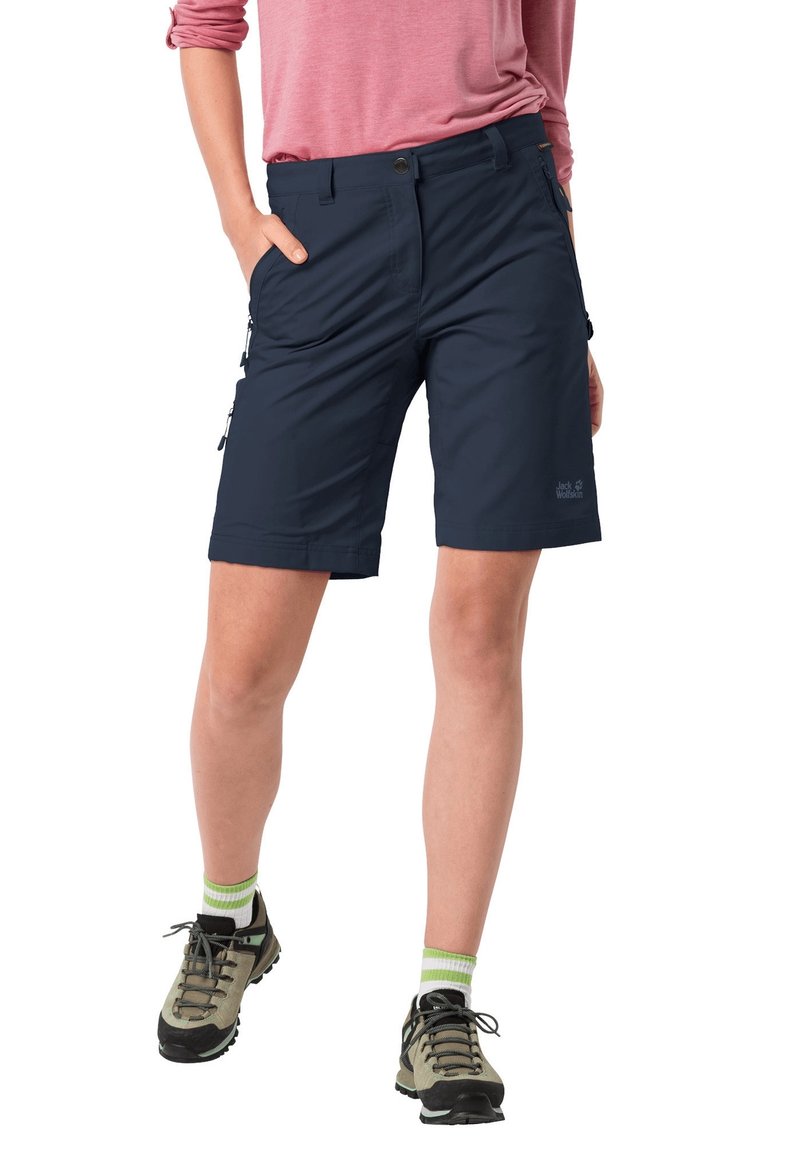 Námořnické modré venkovní kraťasy s volným střihem, zipovými kapsami a odolnou tkaninou. V kombinaci se šedými turistickými botami a ponožkami s zelenými akcenty.